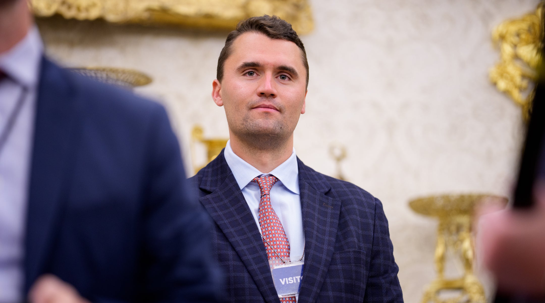 A photo of charlie kirk in a suit wearing a "visitor" badge in the Oval office.