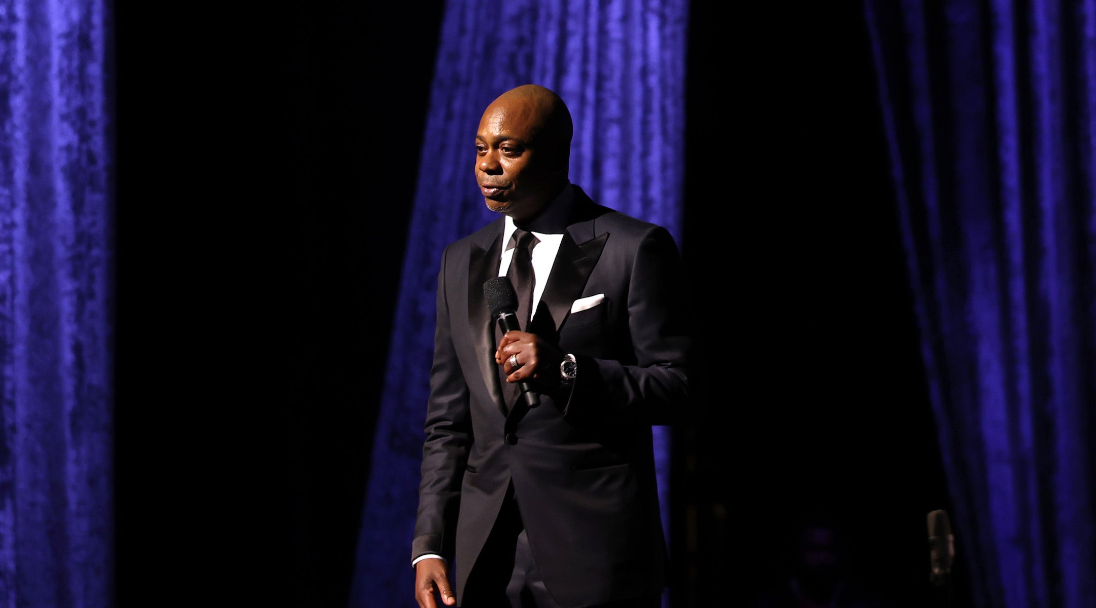 A photo of Dave Chapelle in a suit holding a mic.